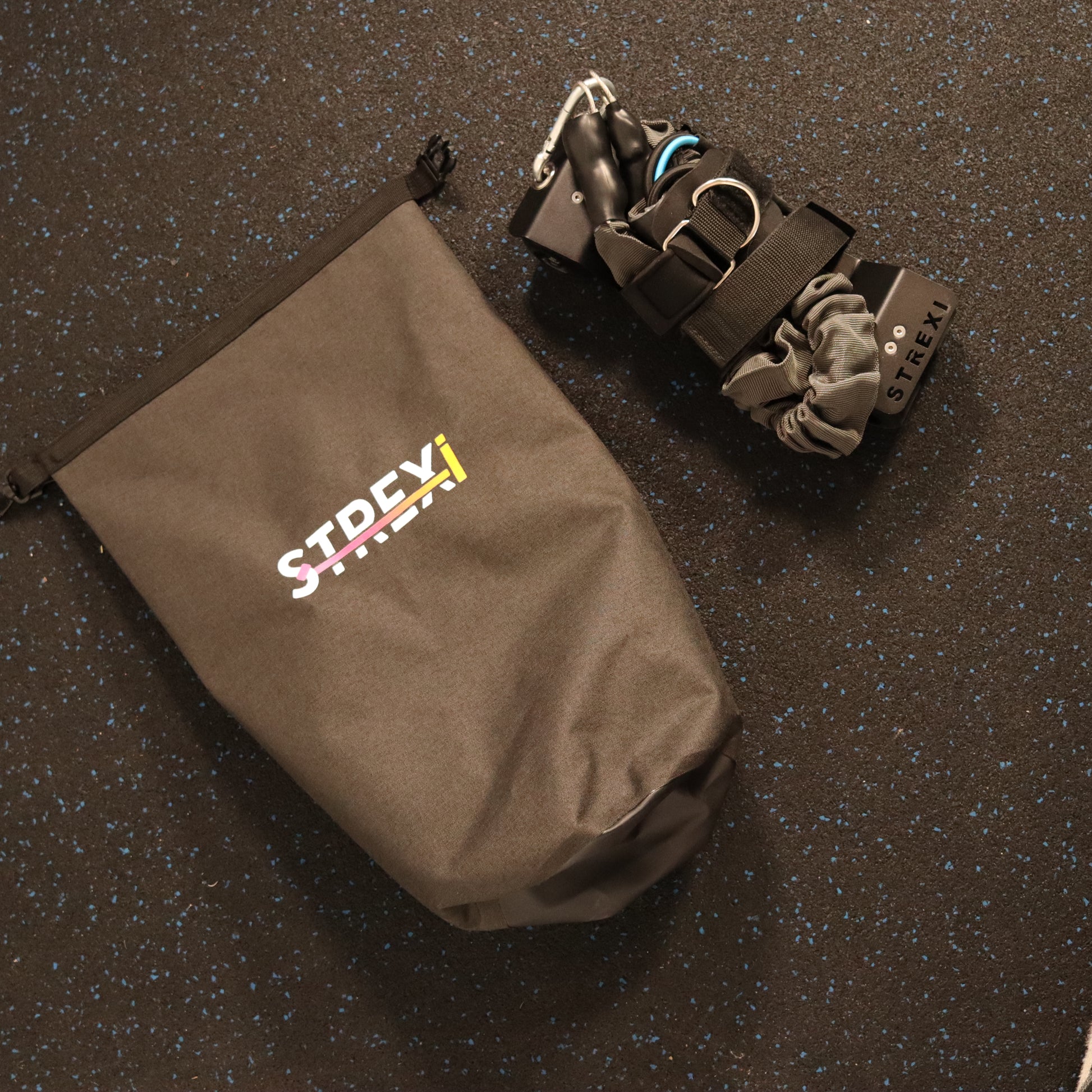 A stretching device named Strexi, along with its grey adjustable safety harness, bands, ankle straps, and a clip shut mechanism, displayed on a carry bag with the Strexi logo visible.
