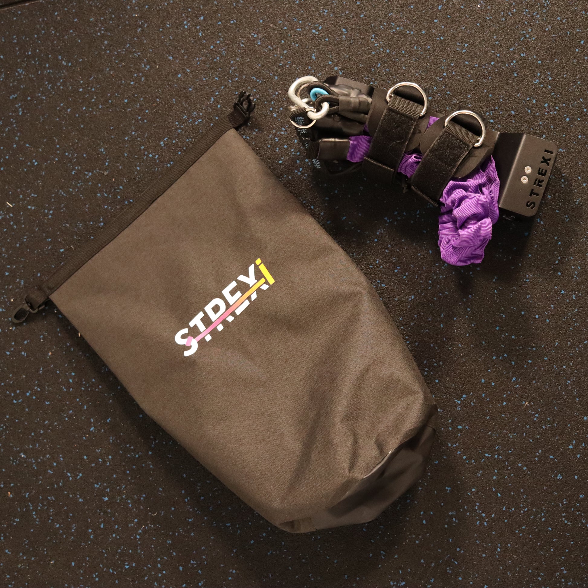 A stretching device named Strexi, along with its purple adjustable safety harness, bands, ankle straps, and a clip shut mechanism, displayed on a carry bag with the Strexi logo visible.